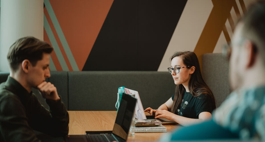 Students working at laptops on a desk, with bold colours striped on the wall behind