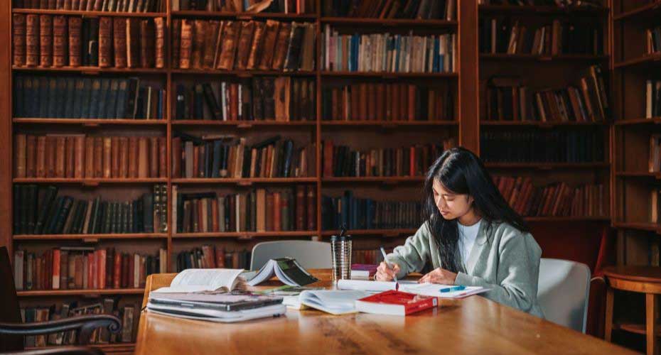 Female students sitting at a desk in the library writing.