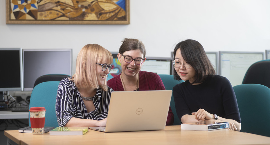 Three women engaged in discussion at a table, with a laptop open in front of them, collaborating on a project.