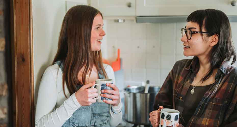 Two female students stood in kitchen facing each other talking whilst holding coffee mugs