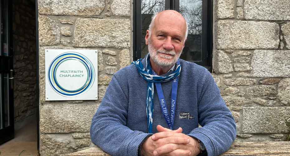 Image of male with white beard smiling at camera with hands clasped, sat outside the multifaith chaplaincy in Cornwall