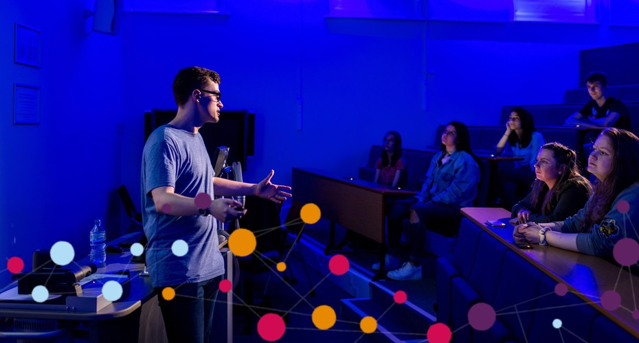 A person giving a presentation with a projector in a blue-lit classroom. Seated audience listens attentively. Overlay of coloured dots and connecting lines on the image.