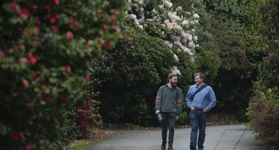 Two pgr students walking along a path in a park in penryn, surrounded by greenery and trees.