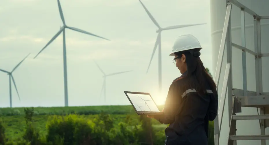 A Lady wearing a white hardhat holding a tablet and looking at a row of wind turbines.