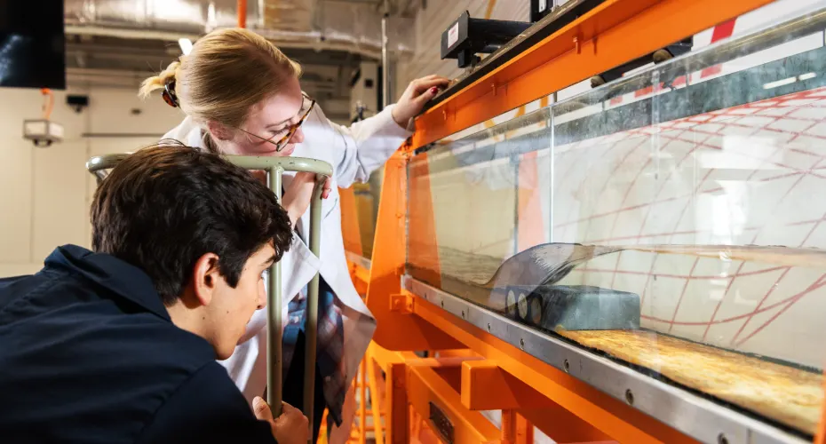 Two students collaborate on a project in a bright orange lab surrounded by various tools and materials for their work.
