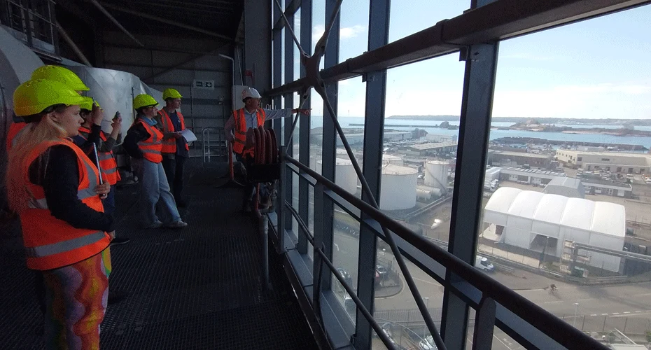 A group of students wearing hi-vis clothing, looking through a window at a large-scale renewable energy site